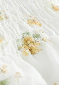 Tissu blanc froncé avec une broderie florale délicate en jaune et vert, créant un aspect texturé et doux.