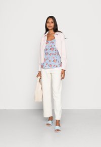 Casaco rosa claro sobre uma blusa peplum floral azul, calças brancas de perna larga e sandálias azuis de deslizar. Segurando uma bolsa bege.
