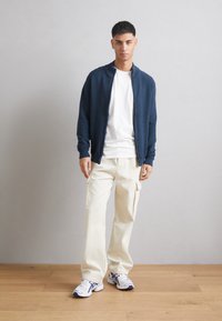 Cárdigan de cremallera azul marino sobre una camiseta blanca, combinado con pantalones cargo crema y zapatillas blancas con detalles en azul. Diseño simple y moderno.