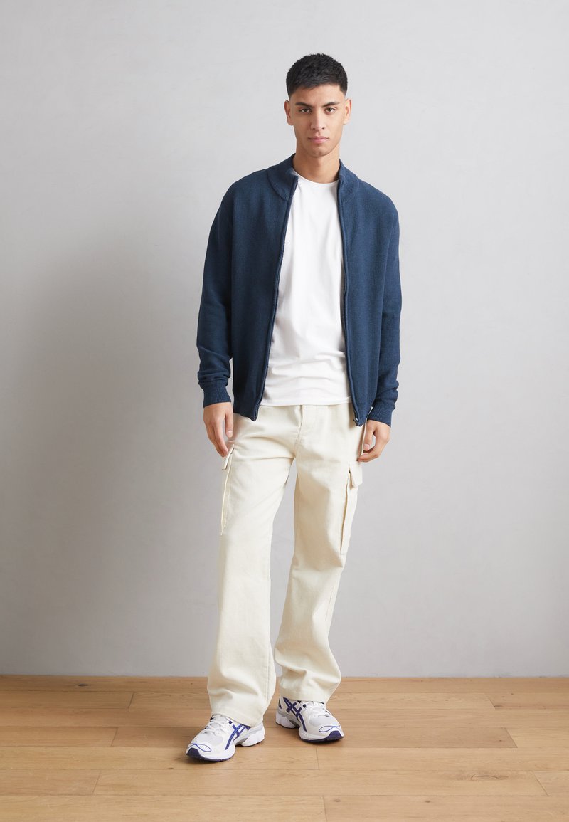 Granatowy sweter z zamkiem błyskawicznym na biały t-shirt, połączony z kremowymi spodniami cargo i białymi trampkami z niebieskimi akcentami. Prosty, nowoczesny design.