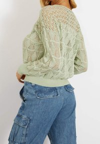 Lichtgroene gebreide trui met een open structuur en een bladvormpatroon, voorzien van een ribbelzoom. Gecombineerd met vervaagde blauwe denim cargo-broeken.