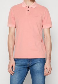 Chemise polo rose clair en coton doux. Dotée d'un col classique, de deux boutons noirs et de manches courtes côtelées. Légère visibilité de la marque.