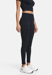 Czarne legginsy z wysokim stanem wykonane z rozciągliwej tkaniny, dopasowany krój o gładkiej fakturze, zestawione z białymi butami sportowymi.
