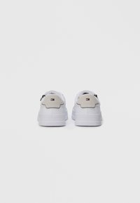 Et par hvite sneakers med beige suedheiskapper og Tommy Hilfiger-logo bak, sett fra bakgrunnen på en ensfarget bakgrunn.