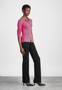 FUKSIA ribatid kardigan kraevi ja nupuvõtuga, koos mustade kohandatud pükste ja mustade terava ninaga kingadega neutraalses taustas.