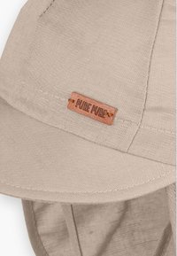 Beige színű pamut sapka varrott peremmel és "PURE PURE" feliratú barna fa címkével. Puha tapintású, és állítható szövetpántokkal rendelkezik.