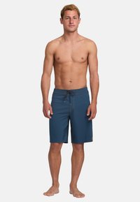Pantaloni da bagno blu con vita regolabile tramite cordino, texture liscia, taglio fino al ginocchio e dettagli di cucitura a contrasto. Adatti per attività acquatiche.