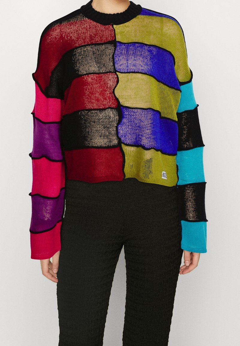 Personne portant un pull tricoté coloré et semi-transparent avec des rayures horizontales rouges, violettes, vertes, bleues et noires, associé à un pantalon noir.