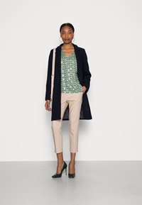 Navy coat, patterned green top with triangles, beige cropped pants, and green high heels. Model holds a light pink bag.