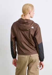 Rudas lengvas švarkas su kapišonu, juodais rankovėmis ir logotipu ant nugaros, dėvimas su khaki spalvos kargo kelnėmis.