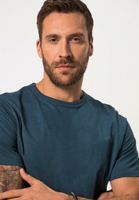 Tyrkysové tričko s kulatým výstřihem, vyrobené z měkké tkaniny, s krátkými rukávy a volným střihem, je nošeno osobou s tetováním na ruce.