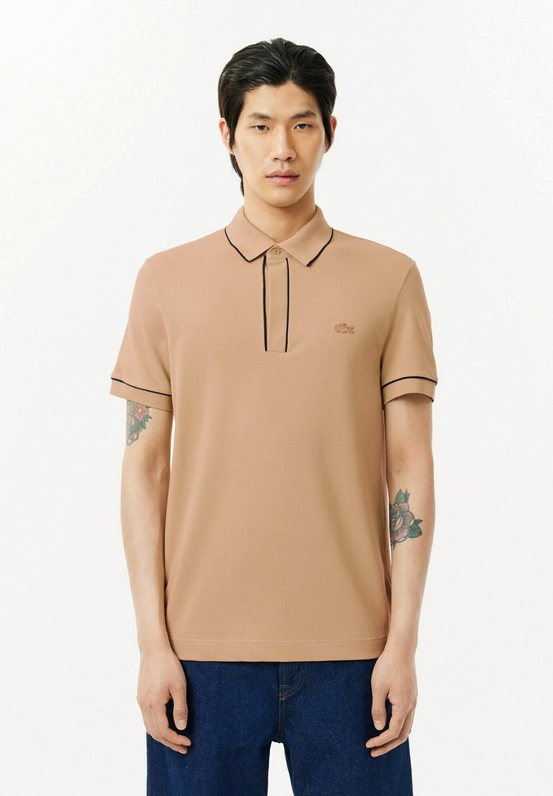 Beżowa koszulka polo z kołnierzykiem, krótkimi rękawami i czarnymi akcentami. Posiada krótki zapięcie na guziki oraz minimalne haftowane logo.