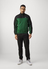 Jachetă sportivă verde și neagră cu fermoar frontale, manșete elastice și pantaloni negri asortați, având un logo în colțul din stânga sus.