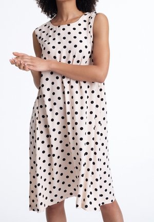 Vrouw draagt een mouwloze beige jurk tot op de knie met zwarte stippen, staat met handen gekruist voor zich tegen een witte achtergrond.