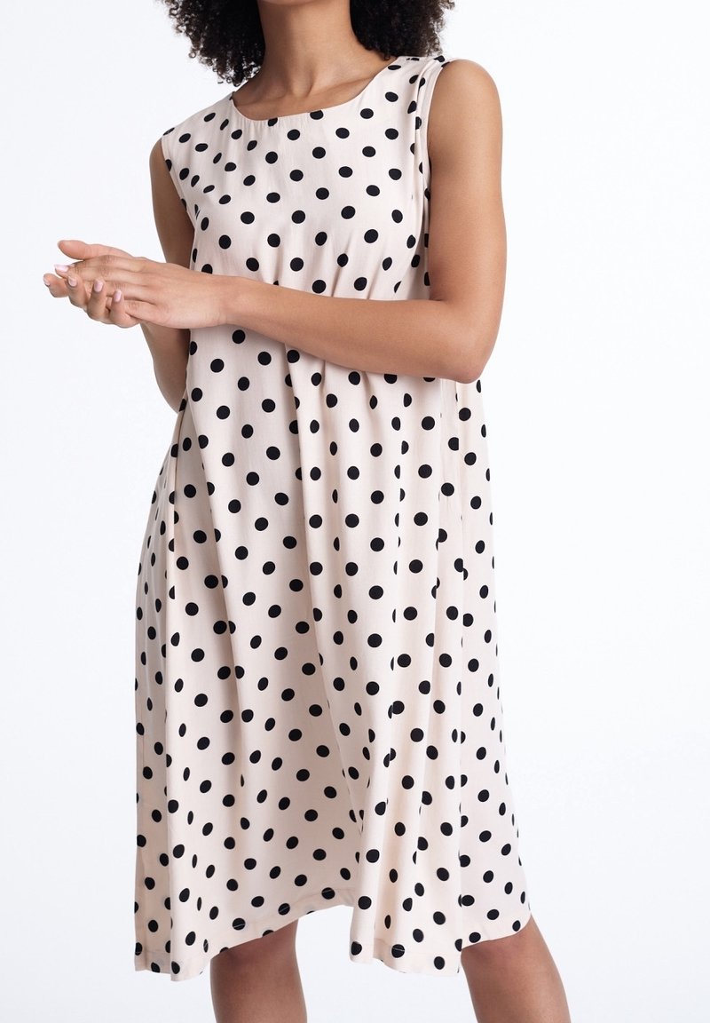 Femme portant une robe beige sans manches, longueur genou, à pois noirs, se tenant debout les mains jointes devant un fond blanc.