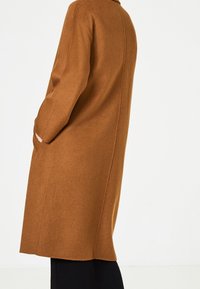 Cappotto di lana marrone con taglio dritto, design minimalista, tasche laterali e colletto alto. Texture morbida e senza chiusure visibili.