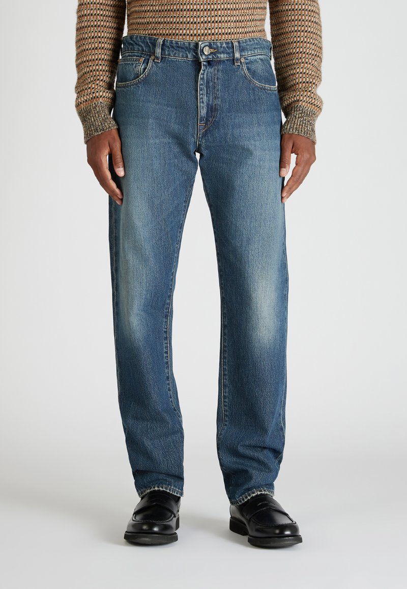 Persona con jeans rectos azules, zapatos negros y un suéter de punto marrón con patrón, de pie frente a un fondo liso.