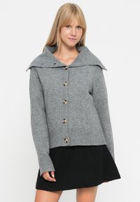Cardigan en laine grise avec un large col, des poignets côtelés et huit grands boutons sur le devant. Texture douce et design légèrement court.