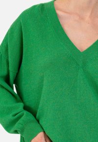 Pull vert à col en V porté par une personne, montrant l'encolure, l'épaule, ainsi qu'une partie d'une manche et de la main.