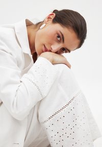 Bela lanena obleka z zapletenim vezenjem in izrezljanimi detajli na rokavih. Model pozira s brado na kolenu in tako prikazuje teksturo tkanine.