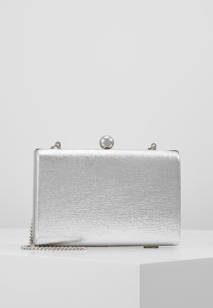 Pochette rectangulaire argentée avec surface texturée, fermoir en perle et fine bandoulière en chaîne reposant sur une surface blanche.