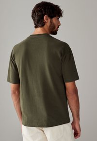 T-shirt in cotone verde oliva visto dalla schiena, con classico scollo a girocollo e maniche corte, texture liscia e orlo dritto.