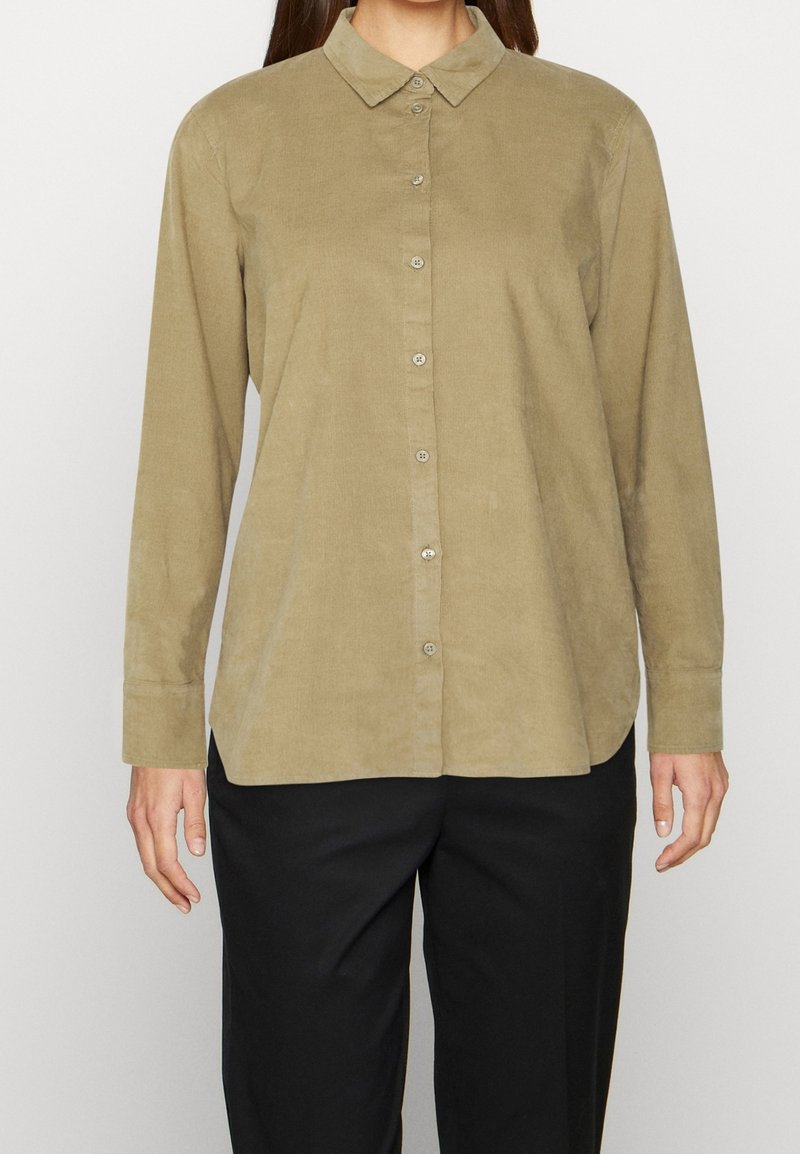 Camisa de pana verde oliva con cierre de botones en el frente, cuello y mangas largas; textura suave y brillo sutil. Pantalones negros no incluidos.
