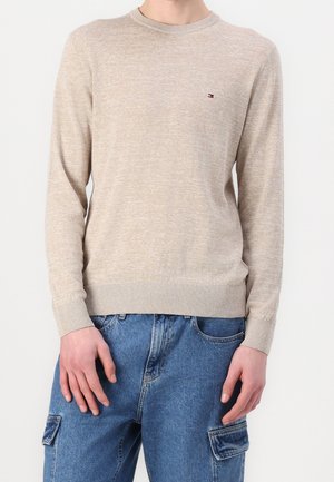 Beige Langarmpullover mit Rundhalsausschnitt, strukturiertem Strick, gerippten Bündchen und Saum. Kleines Logo-Detail auf der Brust. Kombiniert mit blauen Jeans.