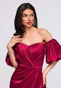 Robe en satin bordeaux à col bateau avec manches bouffantes, corsage ajusté et détails drapés. Portée avec un bracelet argenté et des boucles d'oreilles.