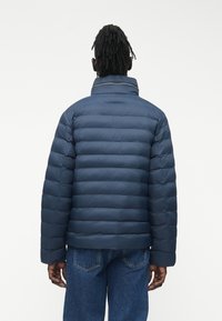 Vīrietis ar drediem, tērpies tumši zilā šūta pūkainā jakā un zilās džinsās, stāvot muguru pret vienkāršu baltu fonu.