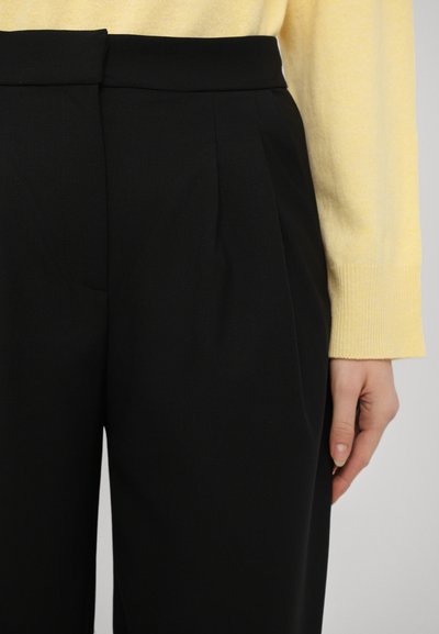 Pantalones negros a medida con pliegues frontales, hechos de una tela suave. Con una cinturilla alta, combinados con un suéter amarillo de manga larga.