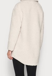 Cappotto oversize in beige chiaro e peloso con una finitura testurizzata, colletto e maniche lunghe. Presenta un'orlo posteriore più lungo e bordi arrotondati.
