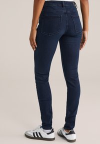 Sötétkék, skinny farmert egy magas derékkal, két hátsó zsebbel és sima denim textúrával, fehér és fekete sportcipővel párosítva.