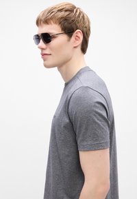 Γκρι κοντομάνικο μπλουζάκι με στρογγυλή λαιμόκοψη, λεία υφή και κεντημένο λογότυπο στο μπροστινό μέρος. Τα αξεσουάρ περιλαμβάνουν μαύρα γυαλιά ηλίου.