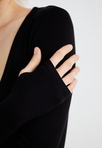 Svart langermet topp med dyp V-hals og tettsittende ermer. Stoffet ser glatt og mykt ut, med en svak glans på overflaten.
