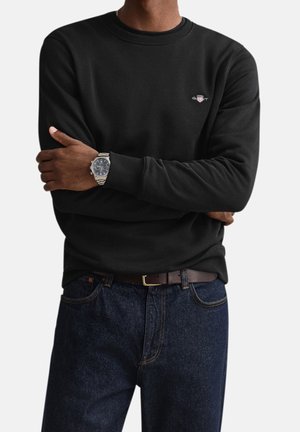 Man met over elkaar geslagen armen, gekleed in een zwart sweatshirt met een klein logo, donkerblauwe spijkerbroek, bruine riem en een zilveren polshorloge met zwarte wijzerplaat.