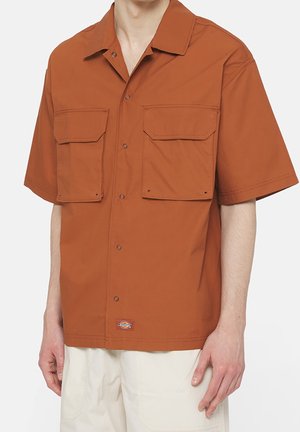 Man draagt een roestkleurig overhemd met korte mouwen met twee grote voorzakken en een beige broek, staand voor een effen achtergrond.