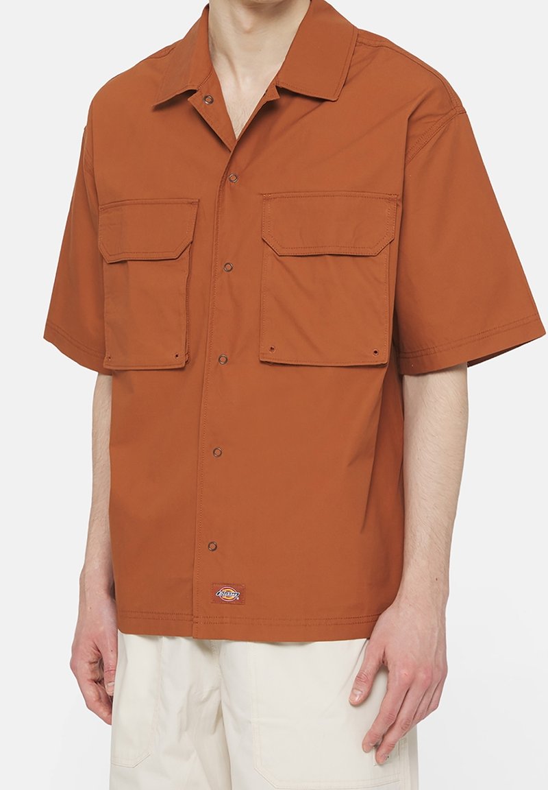 Homme portant une chemise à manches courtes couleur rouille avec deux grandes poches avant et un pantalon beige, debout devant un fond uni.