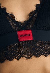 Svart blonde bralette med bølget kant, med et rødt stoffbånd merket "HUGO" og en glatt tekstur.