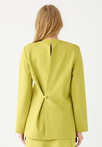 Limegroene blazer met een uitsnijding aan de achterkant en een knoopdetail, op maat gesneden pasvorm en een soepele stoftextuur. Lange mouwen met gestructureerd ontwerp.