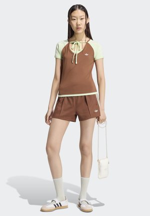 Kobieta ubrana w brązową i jasnozieloną sportową bluzkę oraz szorty, białe siateczkowe skarpetki, białe tenisówki, trzyma białą torebkę z złotym łańcuszkiem.