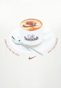 Λευκό φλιτζάνι καφέ και πιατάκι με κουτάλι, ο αφρός καφέ εμφανίζει το λογότυπο "Nike", το κείμενο γύρω αναγράφει "Κατασκευασμένο για τη σόλα" και "Nike sportswear."