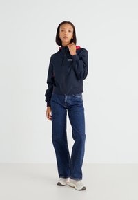 Chaqueta bomber de la marina con acentos rojos, cierre de cremallera, puños de punto y logo frontal. Combinada con pantalones de mezclilla acampanados y zapatillas deportivas.