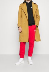 Tan trenchcoat med store knapper, sort hættetrøje, røde sweatpants og hvide sneakers. Glat stof med klassisk snit og tætsiddende manchetter.
