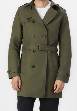 Mężczyzna ubrany w oliwkowo zielony płaszcz dwurzędowy z paskiem w talii i zapinanymi mankietami na białą koszulę.