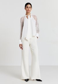 Blusa bianca con maniche in pizzo, scollo annodato e tessuto simile alla seta abbinata a pantaloni bianchi a palazzo e décolleté neri appuntiti.