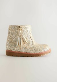 Botín de tobillo dorado con un acabado texturizado, cierre de cremallera en un costado y borlas decorativas. Suela de goma con un tono rosa. Detalle de superficie brillante.
