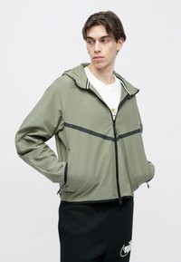 Jasna oliwkowo-zielona kurtka z kapturem, z czarnym zamkiem błyskawicznym i poziomymi czarnymi paskami na wysokości klatki piersiowej. Gładki, lekki materiał.