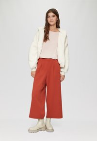 Weiße Plüsch-Jacke mit Reißverschluss, hellrosa Oberteil und weite rostfarbene Hosen. Cremefarbene Stiefeletten mit strukturierten Sohlen. Einfaches Design, keine Muster.