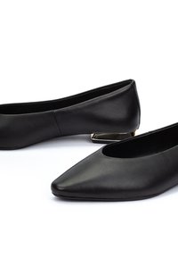 Zapatos planos de punta afilada de cuero negro con un bajo tacón geométrico metálico sobre un fondo blanco.